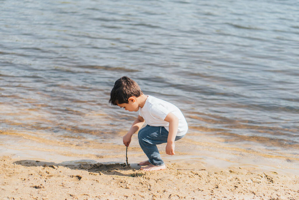 Çıplak ayaklı çocuk ıslak kum ve göletin yanında sopa tutuyor.  - Fotoğraf, Görsel