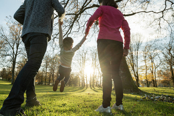 Widok z tyłu młodych rodziców trzymających swoich synów za ręce, podnoszących go w zabawnym obrazie, jak przechadzają się przez park podświetlony słońcem. - Zdjęcie, obraz