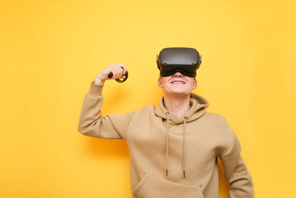 Happy VR gamer winner stands on yellow background, smiles and shows biceps. Portrait of a guy in a VR helmet smiling isolated on yellow. Virtual Reality Cocnept. Copy space - Fotografie, Obrázek