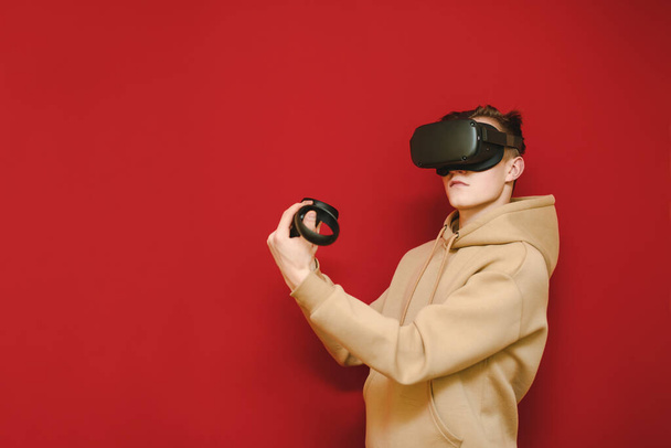 Fotografie mladého muže v zářivé mikině, jak stojí s ovladači v rukou na červeném pozadí a hraje hry na helmě VR, dívá se stranou. Koncept herní přilby VR. Kopírovat prostor - Fotografie, Obrázek