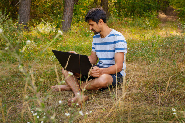 Mladý muž s vousy sedí na trávě a pracuje na počítači. Koncept - na volné noze venku v potěšení - Fotografie, Obrázek