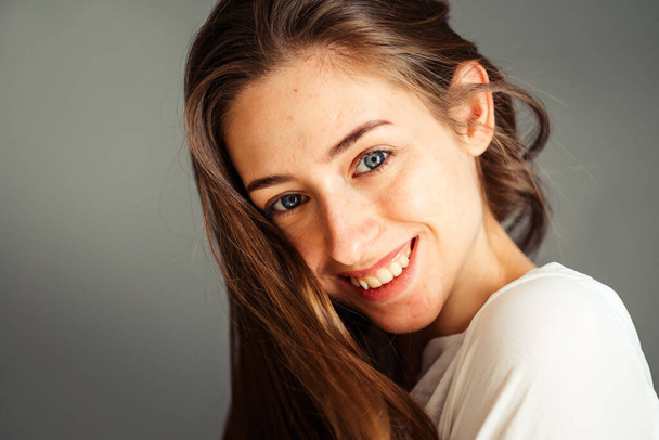 Close-up portret van een lachend jong meisje in een wit shirt op een grijze achtergrond. Handen bij het gezicht. zonder retoucheren en make-up. - Foto, afbeelding