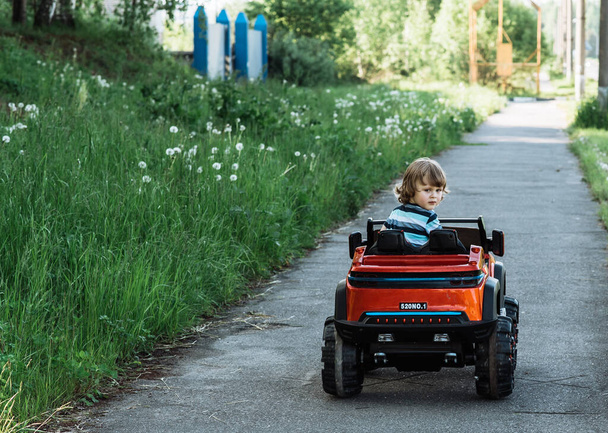 menino de cabelos encaracolados em uma camiseta listrada monta um carro de brinquedo vermelho grande dirigindo em um caminho de asfalto. dia de folga, recreação ao ar livre
 - Foto, Imagem