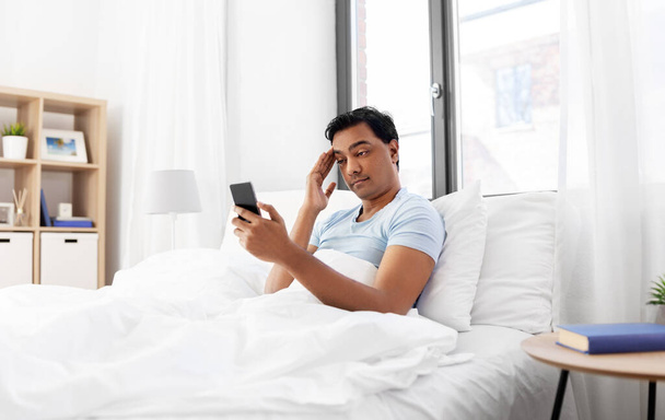 sleepy indian man in bed looking at smartphone - 写真・画像