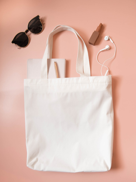 Mock up design bag concept. Top view of blank white tote bag canvas fabric w/ sunglasses, lipstick, notebook, pen and headphone on coral background. Empty eco shopping bag. Flat layout. Vertical. - Zdjęcie, obraz