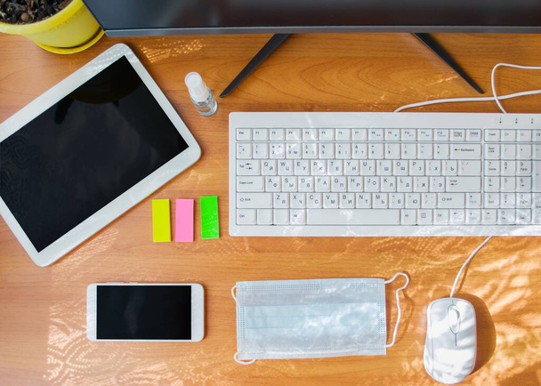 Gadgets, eine weiße Computertastatur und Schreibwaren neben einem Händedesinfektionsmittel und einer medizinischen Schutzmaske auf einem hölzernen Hintergrund. Home Office-Konzept. - Foto, Bild