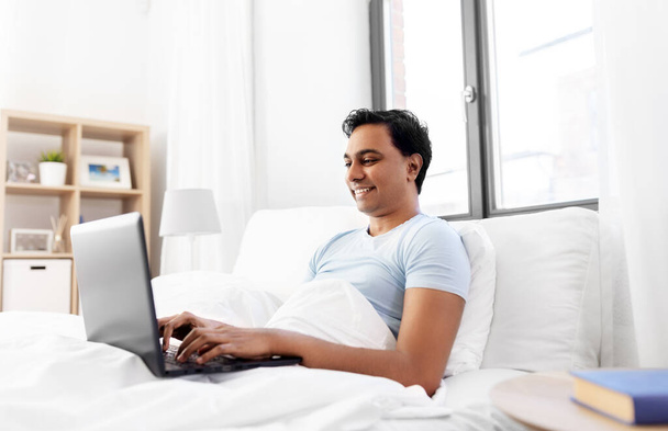 happy indian man with laptop in bed at home - Fotó, kép