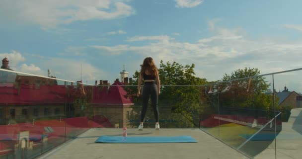 Спортивна дівчина робить присідання зі стрибками. За ним стоїть килимок для йоги та пляшка з водою. Зйомка ззаду і плавний підхід до камери. 4-кілометровий
 - Кадри, відео