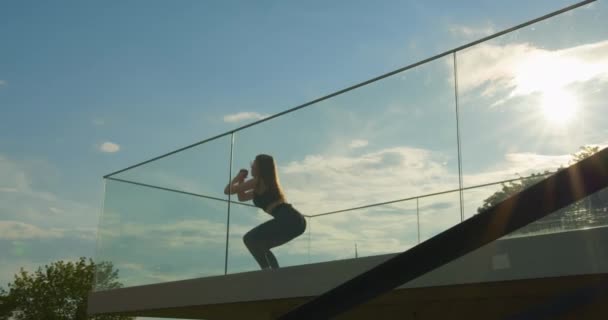 スポーツ少女は飛び降りでスクワットをしています。彼女の髪は飛び跳ねている。サングラスをかけてる。カメラの底から撮影し、滑らかな通路。4K - 映像、動画