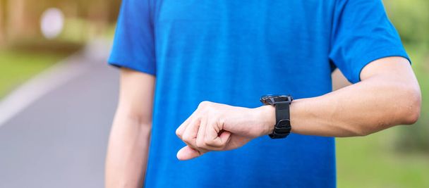 Young adult male checking time and cardio heart rate on sport smartwatch during running in the park outdoor, runner man jogging on the road at morning. Exercise, technology, lifestyle and workout - Foto, Imagen