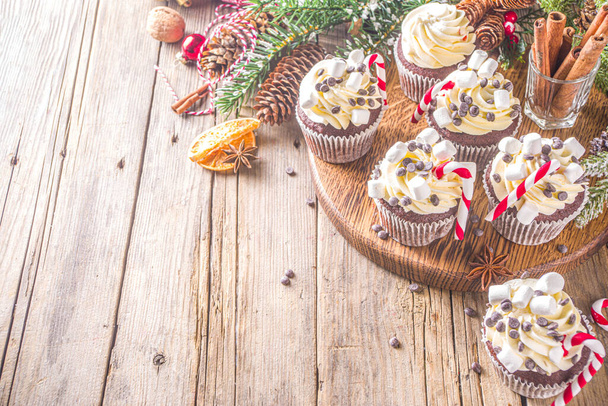 Christmas cupcakes. Xmas hot chocolate cupcake with peppermint candy cane, chocolate drops and marshmallow. Wooden background with Christmas decor and fir tree branches. Sweet Christmas baking/ - 写真・画像