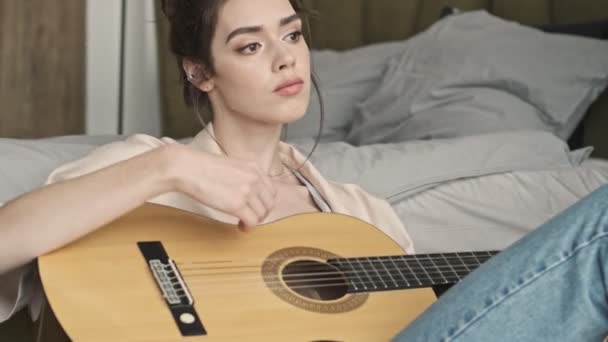 Young beautiful concentrated woman indoors at home sitting near bed while playing on a guitar - Filmmaterial, Video