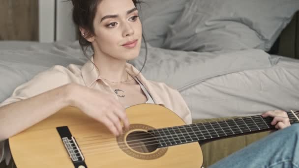 Pretty concentrated woman indoors at home sitting near bed while playing on a guitar - Záběry, video