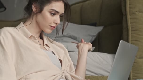 Young pleased woman sitting indoors at home while using laptop computer and writing notes with pencil - Footage, Video