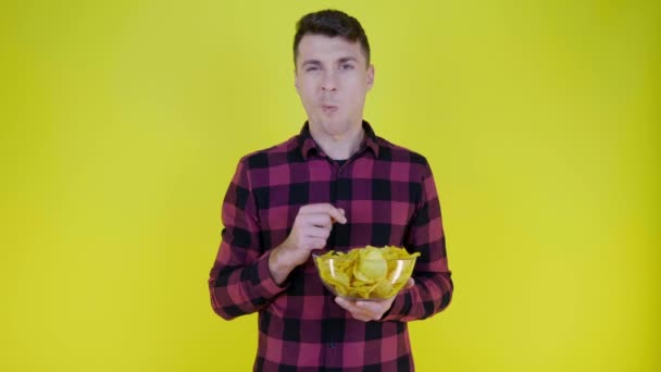 Muž v růžové kostkované košili jí bramborové lupínky se skleněnou miskou na žlutém pozadí - Záběry, video