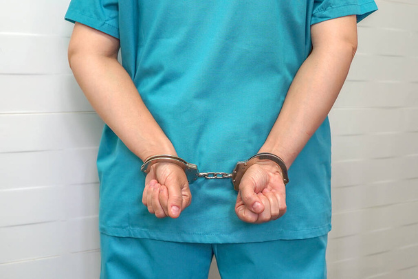 Έγκλημα στην ιατρική, χειρουργός ή γιατρός με μπλε στολή με τα χέρια δεμένα με χειροπέδες στην πλάτη. Εγκληματολόγος γυναίκα χέρια κλειδωμένο σε χειροπέδες. - Φωτογραφία, εικόνα