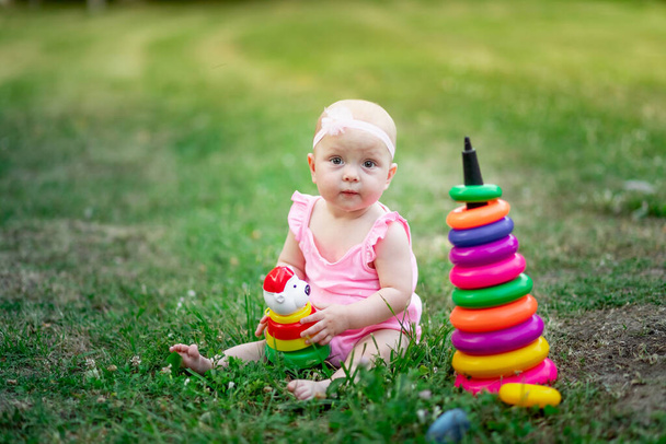 κοριτσάκι 10 μηνών κάθεται στο γρασίδι το καλοκαίρι και παίζει πυραμίδα, πρώιμη ανάπτυξη των παιδιών, υπαίθρια παιχνίδια - Φωτογραφία, εικόνα