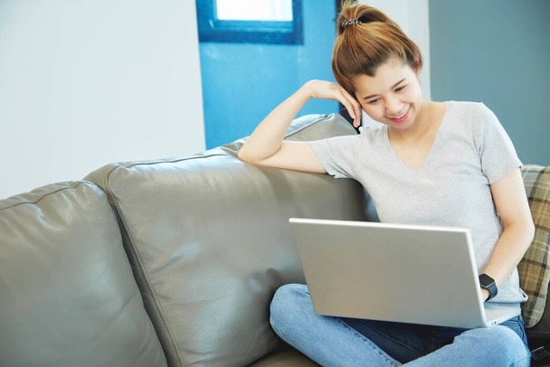 Женщина азиатка сидит на диване, используя ноутбук дома. Работа на дому
 - Фото, изображение