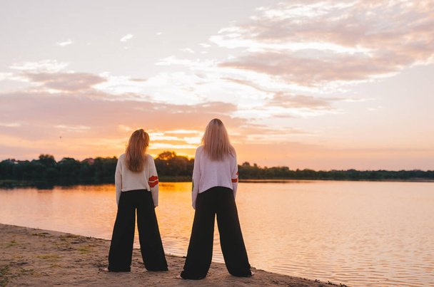 Δύο γυναίκες που κάνουν διαλογισμό γιόγκα την αυγή. Πρωινή γιόγκα στην παραλία ή στην ακτή του ποταμού. Πίσω όψη.  - Φωτογραφία, εικόνα