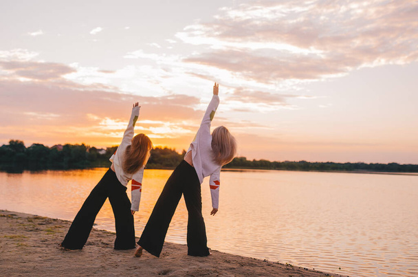 Zwei Frauen praktizieren Yoga-Meditation bei Sonnenaufgang. Morgens Yoga am Strand oder an der Flussküste. Schöne Aussicht.  - Foto, Bild