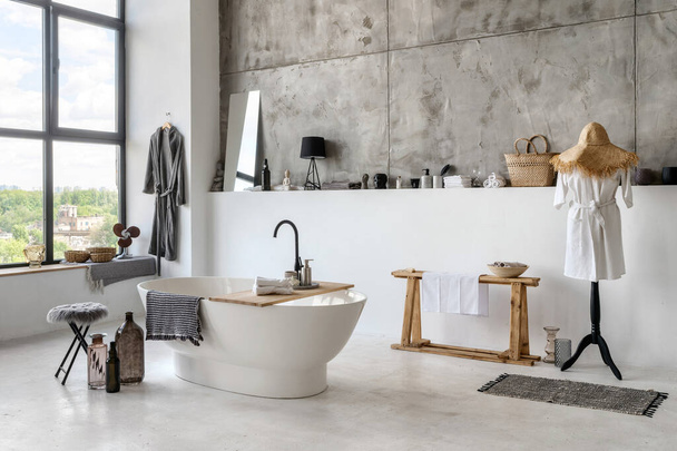 Modern house with contemporary interior design, white bathroom, comfortable bathtub and home decor close to mannequin in cotton bathrobe near wooden bench with clean towels - Fotó, kép