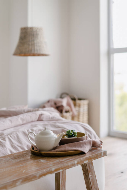 Selective focus on vertical photo of bench with home decor and teapot on tray. Morning in cozy bedroom with crumpled blanket on comfortable bed near laundry basket - Photo, Image