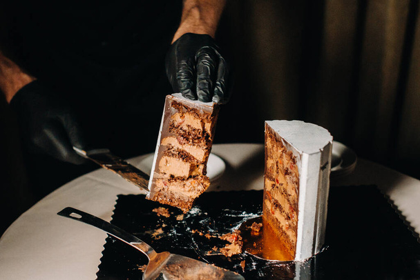 Geweldige Cakes. Een chef-kok met zwarte handschoenen snijdt een chocoladetaart. De bruidstaart is heerlijk binnen op een zwarte achtergrond. Grote taart in witte chocolade - Foto, afbeelding