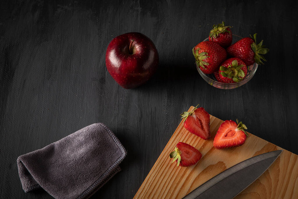 Strawberries in a glass bowl on a wooden table, with a cutting board, with cut strawberries and a knife next to it, accompanied by a red apple. - Foto, imagen