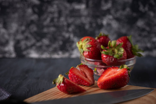 Strawberries in a glass bowl on a wooden table, with a cutting board with cut strawberries and a knife next to it. - 写真・画像