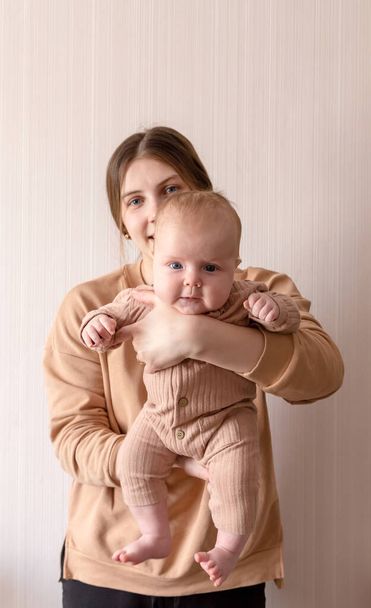 A young mother holds a girl, who is 3 months old, in her arms. Mom and baby in Brown clothes on a light background - Zdjęcie, obraz
