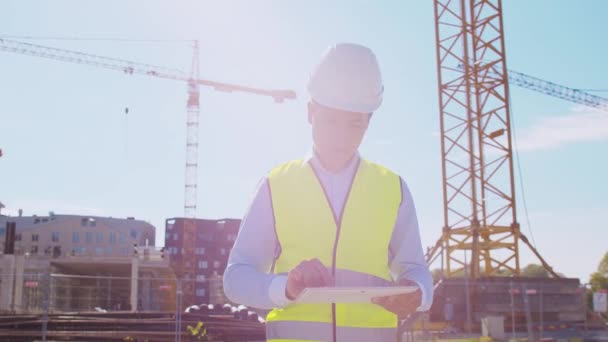 Professional builder standing with tablet computer in front of construction site. Foreman in helmet and vest. Office building and crane background. Business, real estate and investment concepts. - Кадры, видео