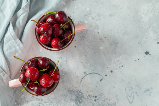 Two pink mugs with fresh ripe cherries. Sweet organic berries on a light concrete background. Top view with copy space - 写真・画像