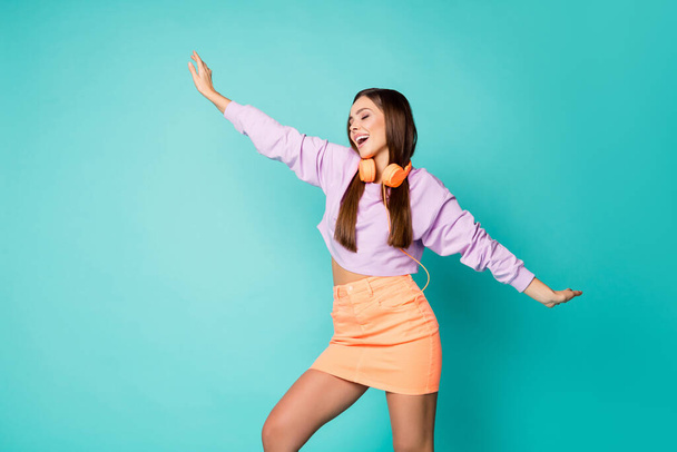 Foto von niedlichen schönen Millennial Dame hören moderne Technologie Ohrhörer tanzen überglücklich heben Hände Jugend bewegt tragen violetten Pullover orange Minirock isoliert teal Farbe Hintergrund - Foto, Bild