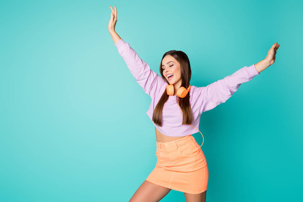 Photo of cute lovely millennial lady listen modern technology earphones dancing overjoyed raise hands youth moves wear cropped violet sweater orange skirt isolated teal color background - Foto, Imagem