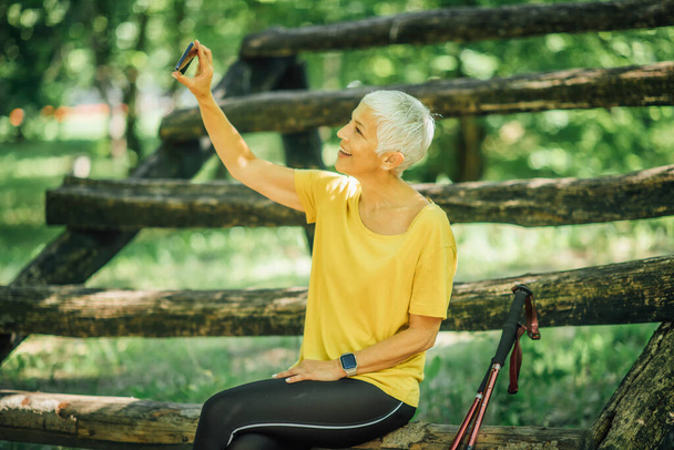 Mulher idosa ativa satisfeita fazendo uma pausa na natureza, tomando selfie após o exercício de caminhada nórdica
 - Foto, Imagem
