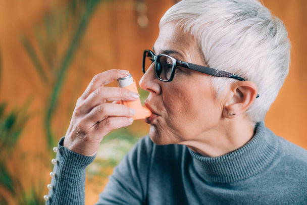 Старшая женщина использует ингалятор астмы с удлинителем трубки
 - Фото, изображение