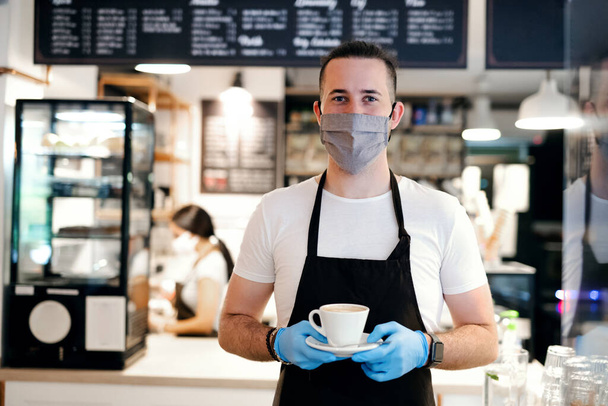 Waiter with face mask and gloves working in cafe, holding coffee. - Photo, image