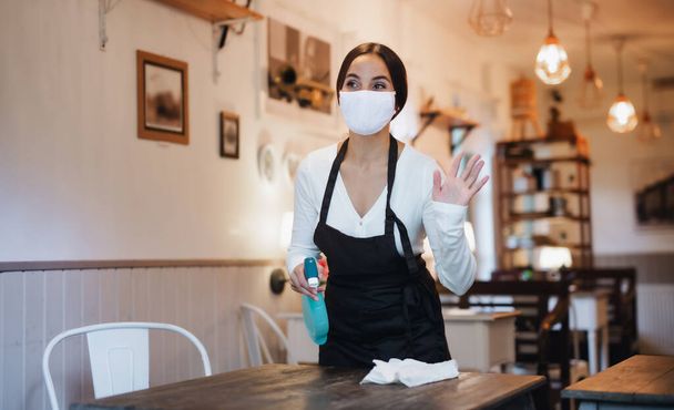 Young woman with face mask working indoors in cafe, disinfecting tables. - Fotografie, Obrázek