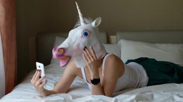 Woman with a head mask of unicorn talking to web camera on smart phone on a bedroom. Social media concept. Weird funny video - Filmagem, Vídeo