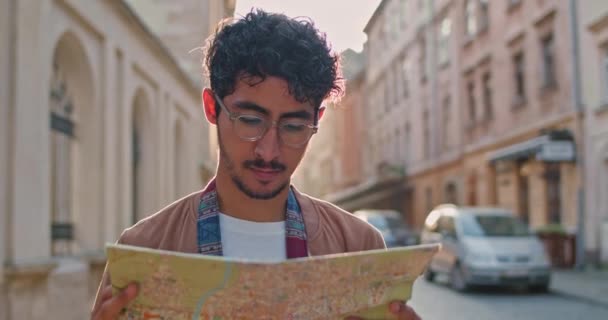 Close up of male tourist looking at map and smiling while standing at old city street. Millennial man in glasses finding direction with map.Concept of travelling and tourism. - Filmagem, Vídeo