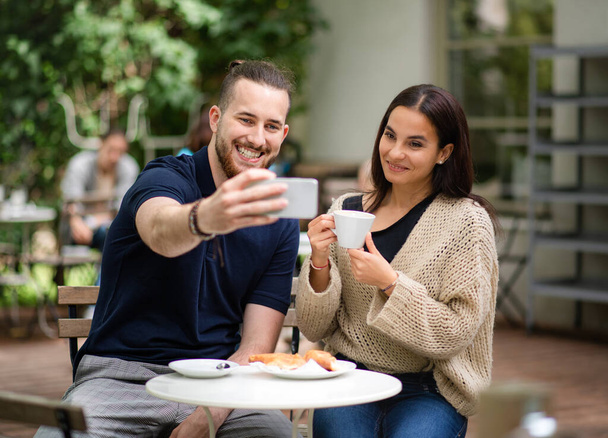 Happy young couple sitting outdoors in cafe, taking selfie by smartphone. - Zdjęcie, obraz