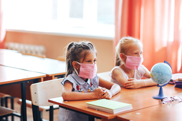 Dwie małe uczennice siedzą przy biurku w klasie szkolnej w maskach medycznych i uważnie słuchają nauczyciela - Zdjęcie, obraz