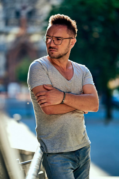 stylish guy with glasses looks away, holds onto the handrail and thinks. strong male gaze. light jeans and a fashionable gray T-shirt. contour sunlight. summer. - Photo, Image