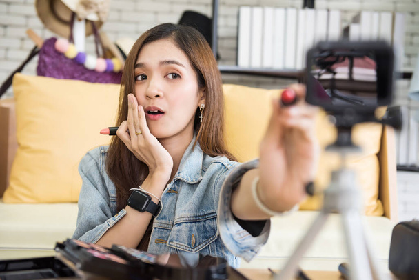 Beauty blogger γυναίκα δείχνουν κραγιόν μακιγιάζ για να ζήσουν streaming βίντεο κάμερα στο τρίποδο στο σπίτι. Ασιατικό χαριτωμένο νεαρό κορίτσι πωλούν καλλυντικά σε απευθείας σύνδεση. internet influencer κινηματογραφώντας καθημερινά make-up ρουτίνα φροντιστήριο. - Φωτογραφία, εικόνα