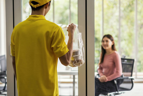 Toimittaja keltainen univormu toimittaa kakku jälkiruoka muovipusseihin hämärtynyt liikenainen asiakas toimistorakennuksessa. Onnellinen tyttö odottaa ruokaa. Elämäntapa työpäivänä. Uusi normaali käsite - Valokuva, kuva