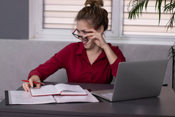 La rubia feliz con camisa roja y gafas rojas trabaja en la oficina o en casa. trabajo remoto o estudio. Con portátil
 - Foto, imagen