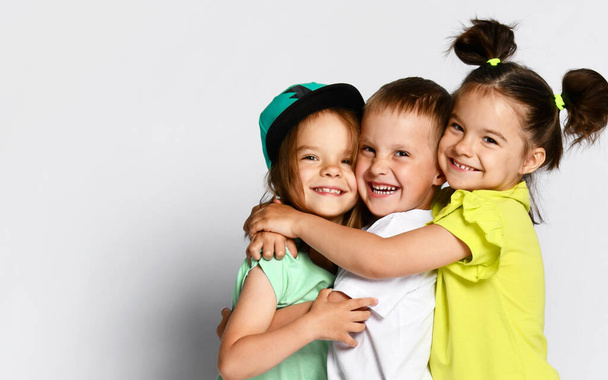 Studio portrait of children on a light background: full body shot of three children in bright clothes, two girls and one boy. Triplets, brother and sisters. hugging on camera. Family ties, friendship - 写真・画像