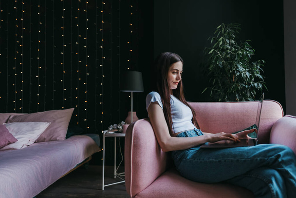 Beautiful young caucasian woman working with laptop computer, sitting on a pink sofa at home near the bed. People work from home. High quality photo - Foto, afbeelding