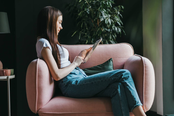 Young caucasian woman sitting on pink sofa and using a tablet computer, typing text, freelance worker . High quality photo - Photo, Image