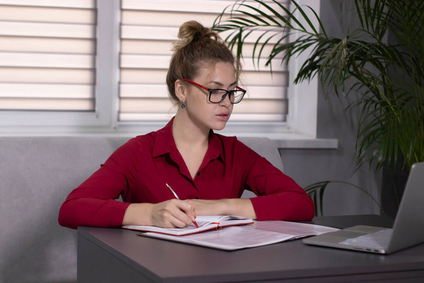 blond in een rood shirt en rode bril werkt op kantoor of thuis. op afstand werken of studeren. Met een grijze laptop. - Foto, afbeelding